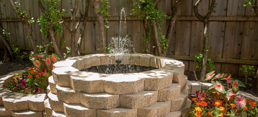 A circular above ground paver pond sits in a flower bed, shaded by the tree. On either side are semi-circular paver flowerbeds. Behind the water feature is a wooden fence and landscaped trees.