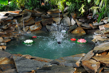 A beautiful pond with blue water. It has a waterfall, fountain, and blue tinted water. There are flat landscaping stones surrounding the pond, and lush green foliage. Floating in the pond are faux lilies.