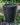 A black planter with a stone finish sits on landscaping stones amongst green plants and flowers. The planter is a disappearing water feature. The fountain water is flowing.